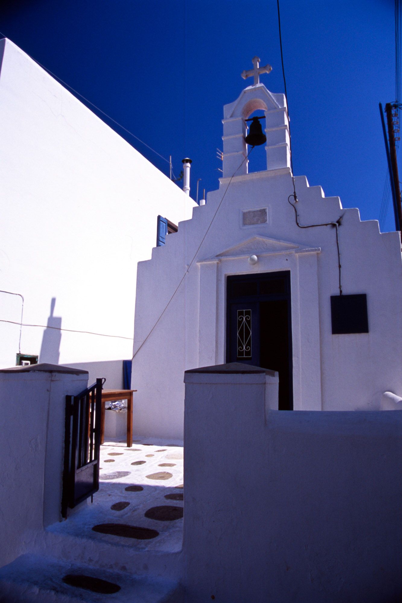 White-washed Greek Orthodox chapel with bell tower and cross against a deep blue sky.