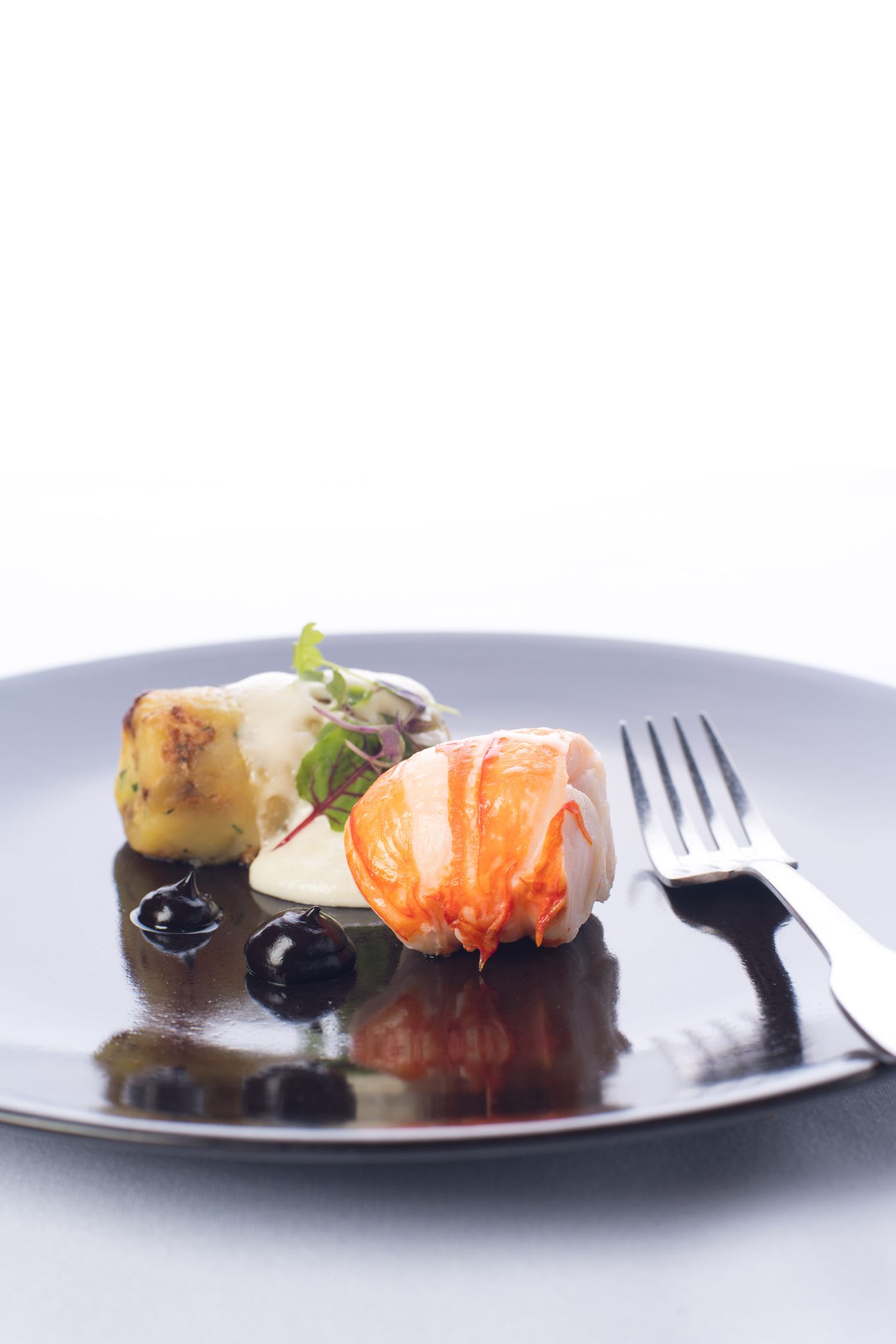 Gourmet seafood dish with lobster, potato and dark sauce on a black plate.