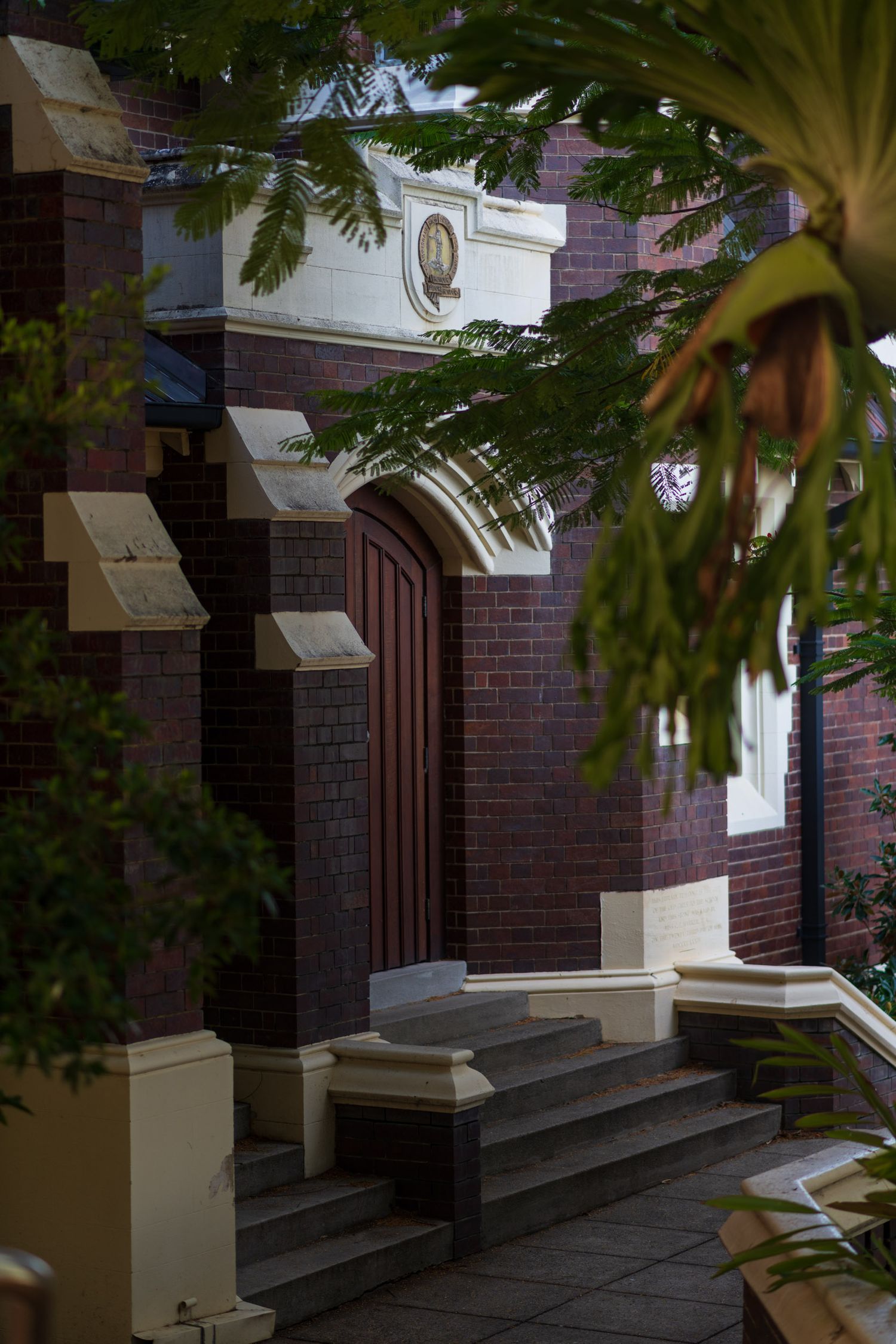 Historic red brick church entrance with arched wooden door, white stone detailing, and lush green trees in Brisbane.