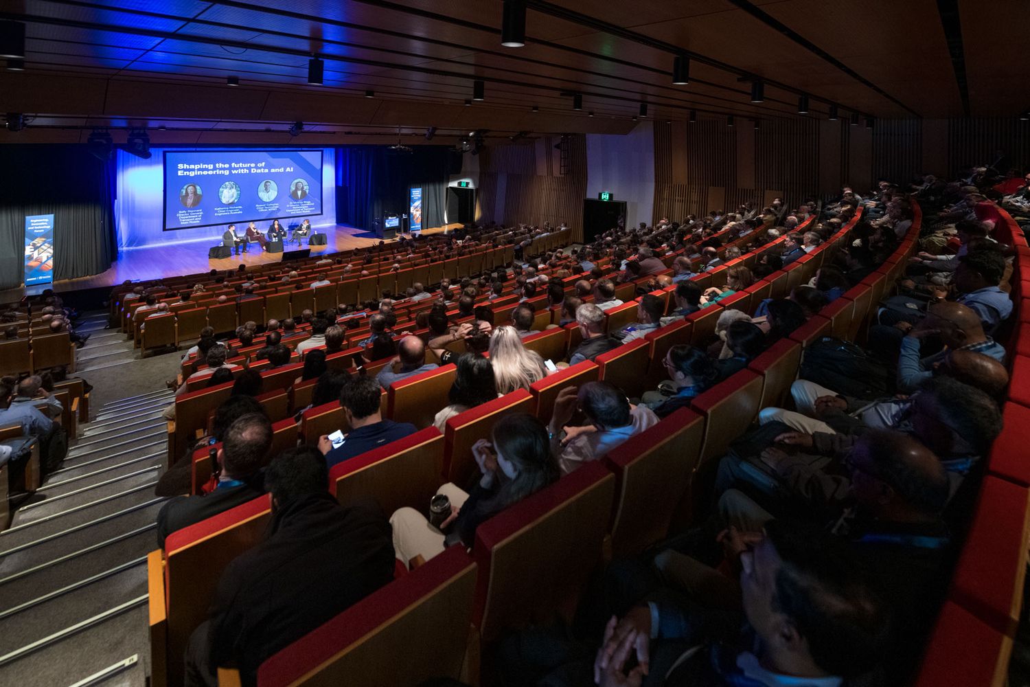 Large conference auditorium filled with attendees watching a panel presentation on a blue-lit stage.