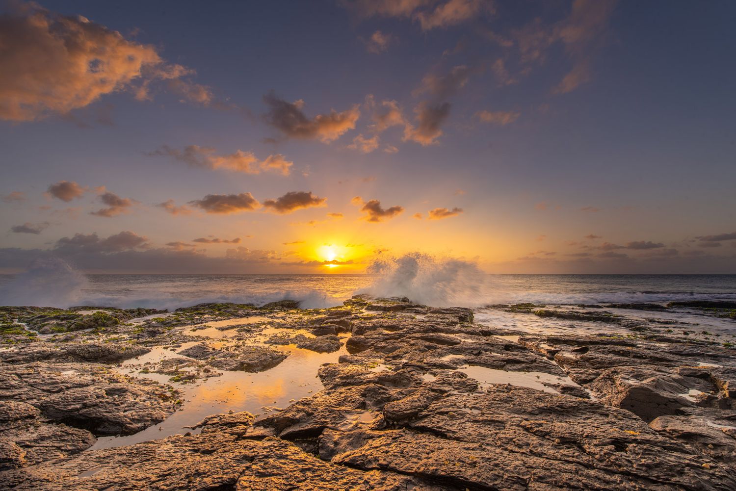 Golden sunset over rocky ocean coastline with crashing waves and colorful clouds reflected in tide pools.