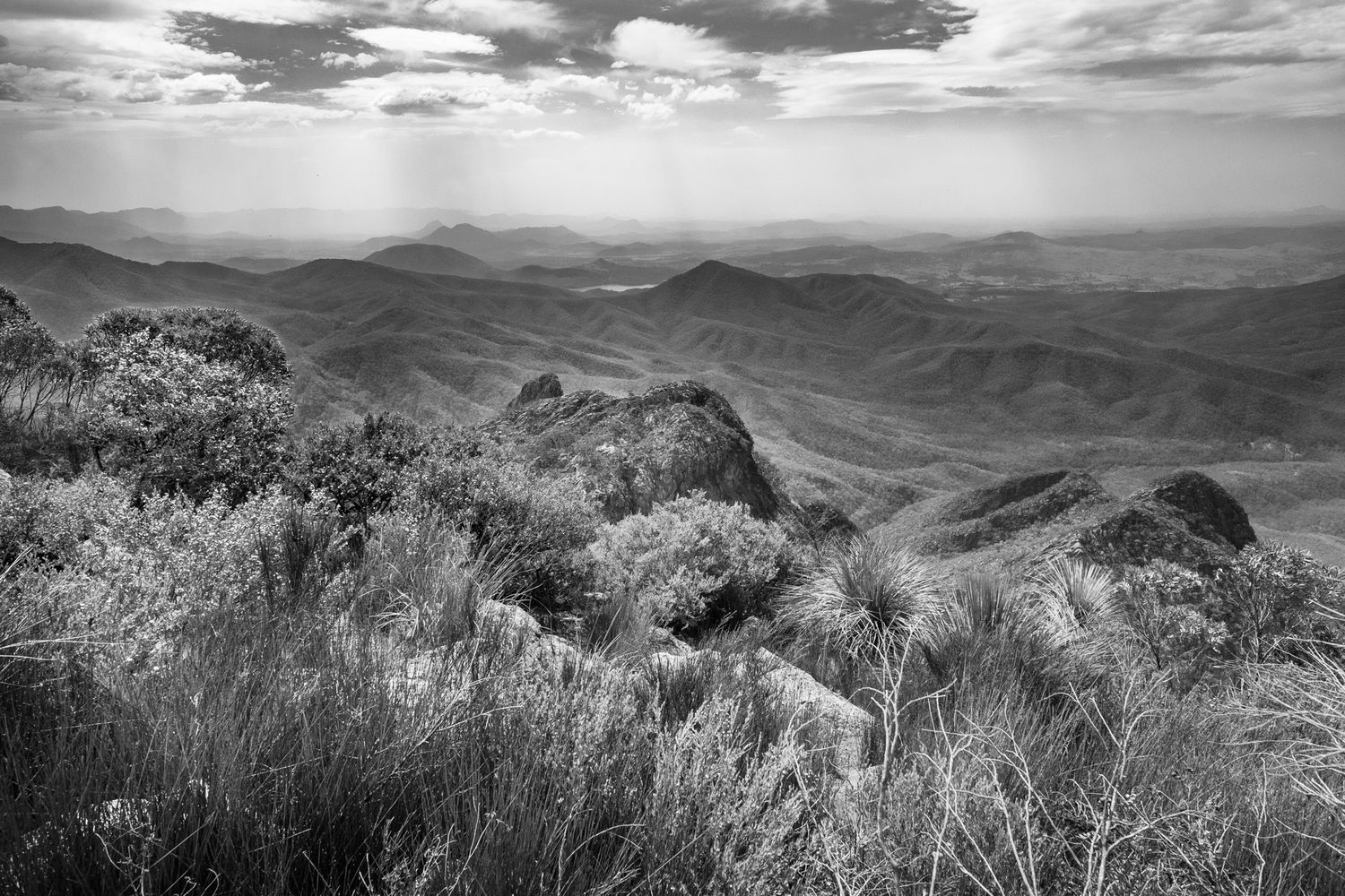 Black and white aerial view of vast mountain ranges with rocky outcrops and shrubland under cloudy skies.