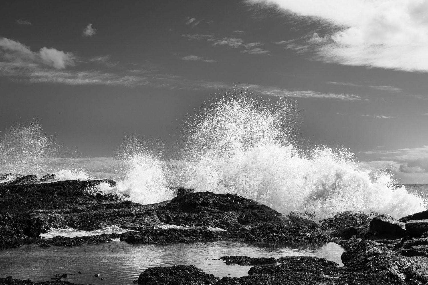 Black and white dramatic ocean wave crashing over rocky coastline under cloudy sky.