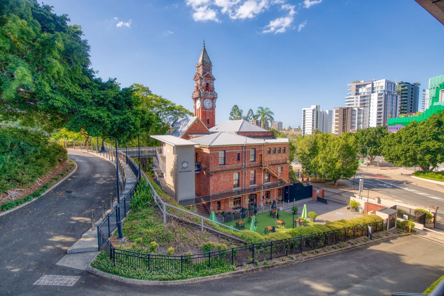 Aerial view of historic red brick church with clock tower surrounded by trees in urban Brisbane cityscape.