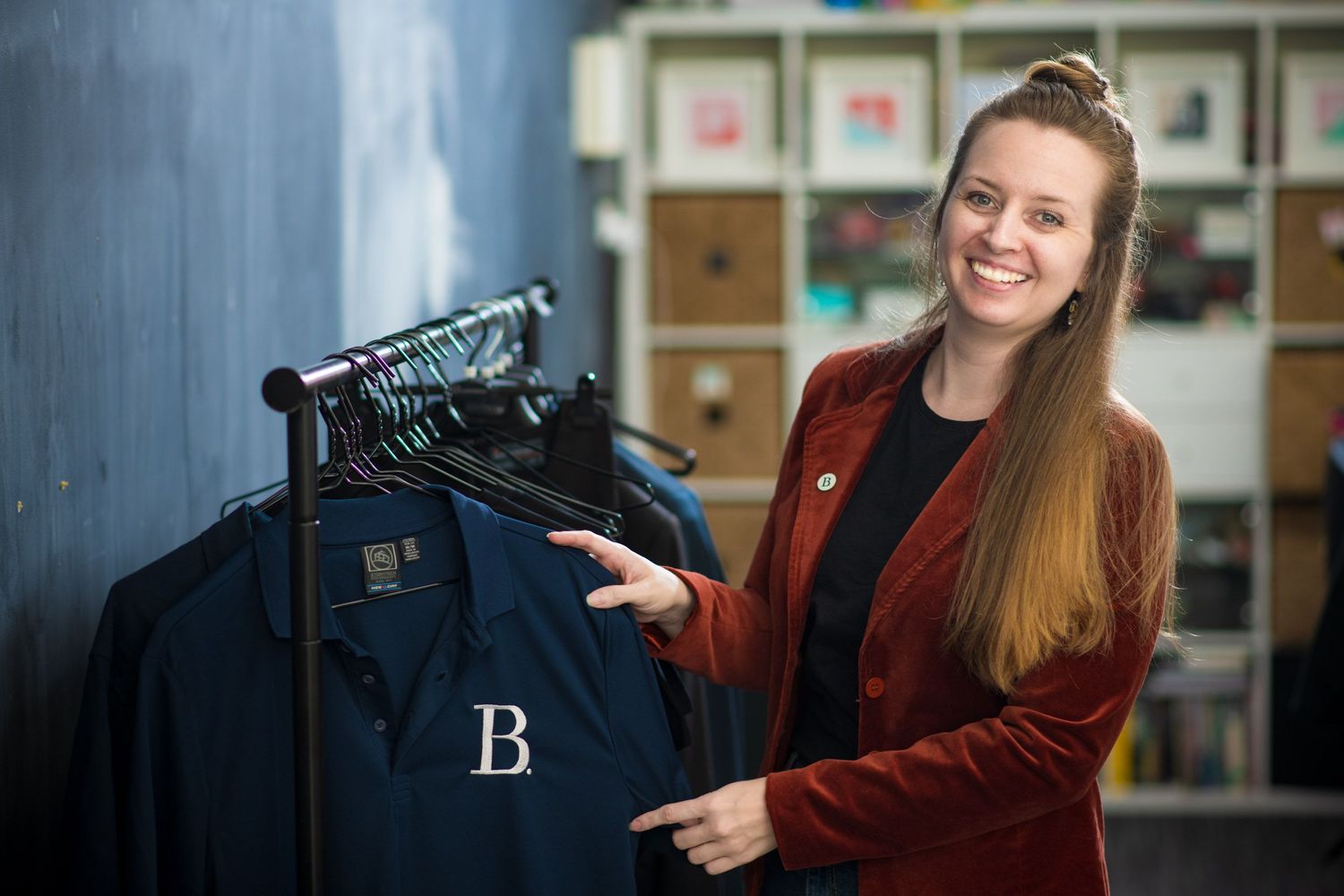 Young woman in red blazer smiling while pointing at branded clothing on a rack in a retail store.