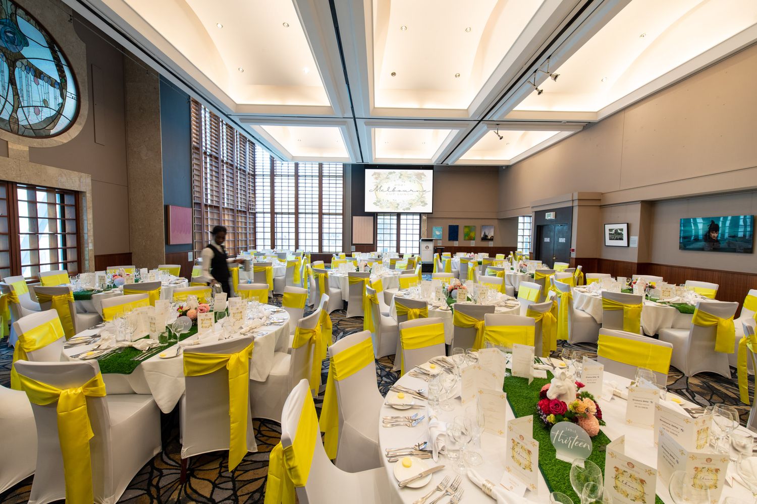 Elegant banquet hall with round tables decorated with yellow chair sashes and green centerpieces for a formal event.