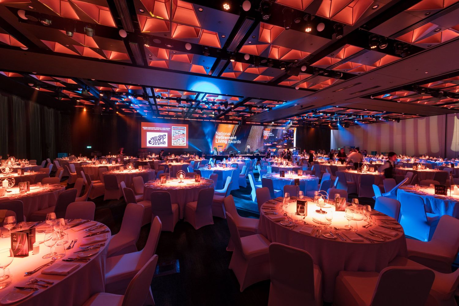 Spacious gala ballroom with warm amber and blue lighting, round white-clothed tables, and projection screens.