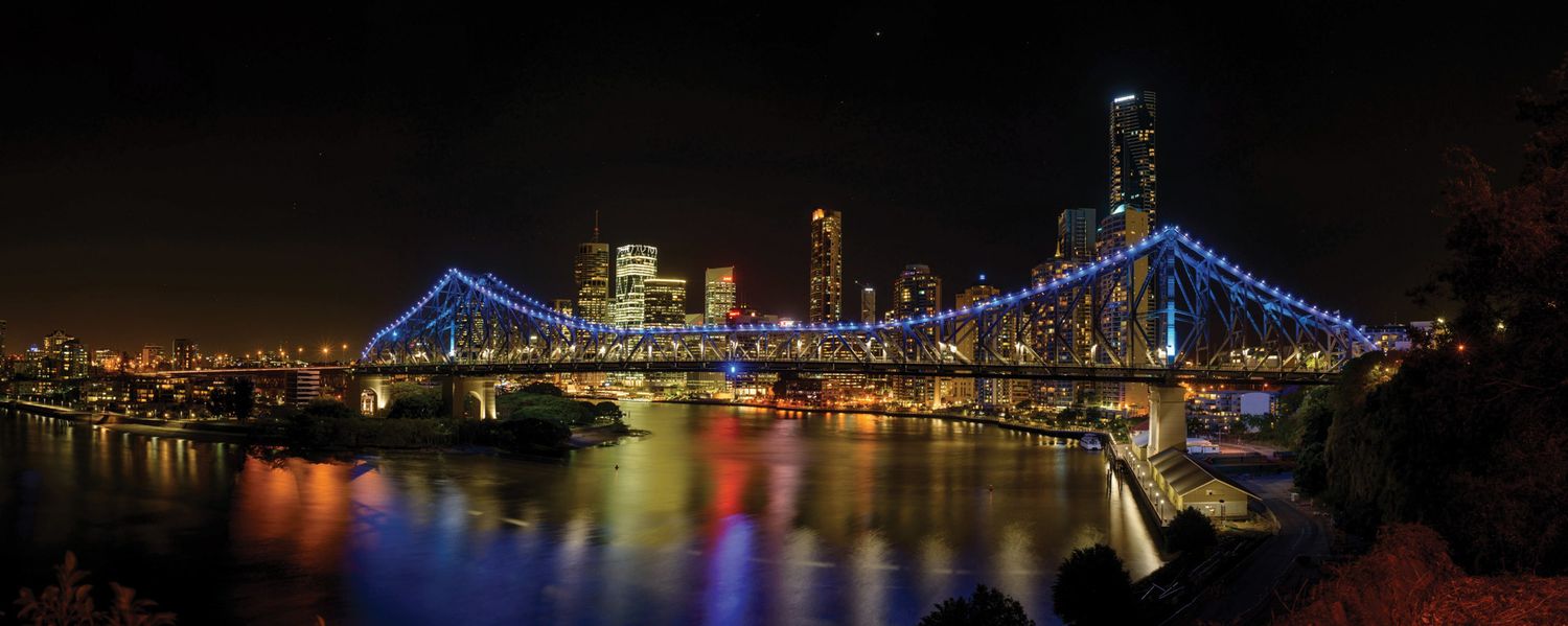 Story Bridge illuminated in blue lights reflecting on Brisbane River at night with city skyline backdrop.