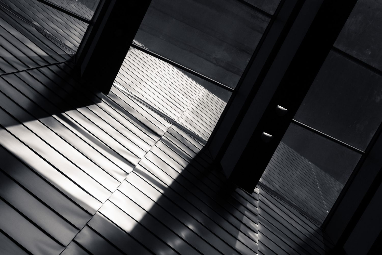 Black and white abstract photo of light streaming through architectural slats onto a wooden floor.