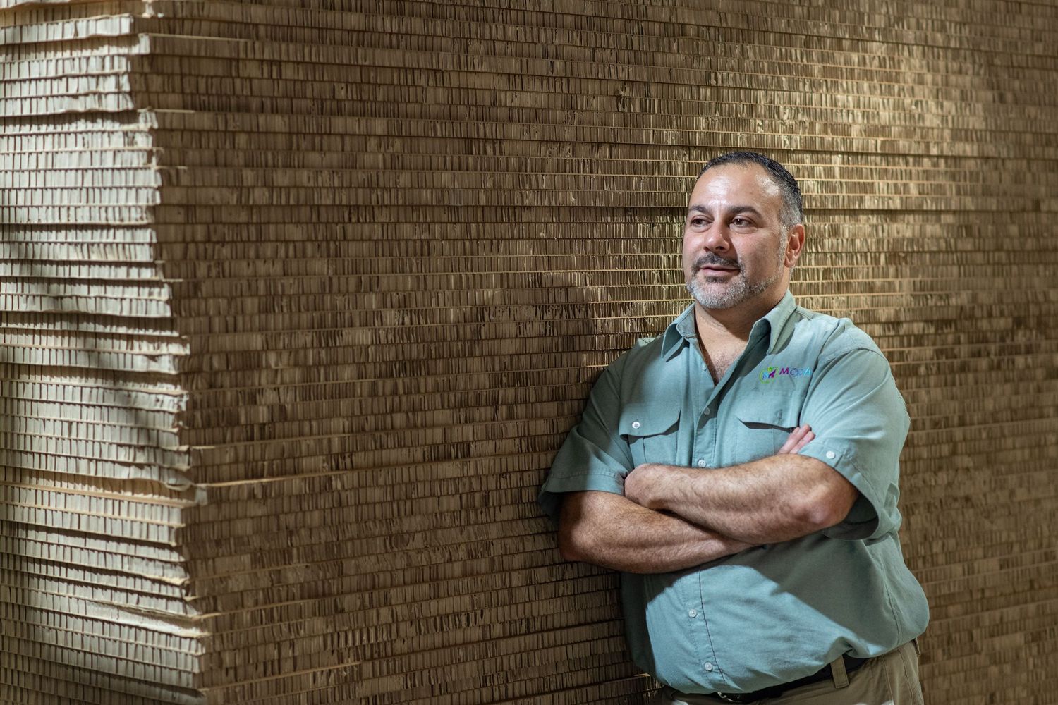 Confident man with crossed arms leaning against a large stack of cardboard in a warehouse.