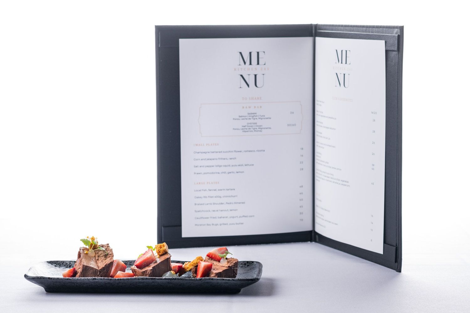 Elegant restaurant menu book displayed open beside a dark plate of gourmet appetizers.