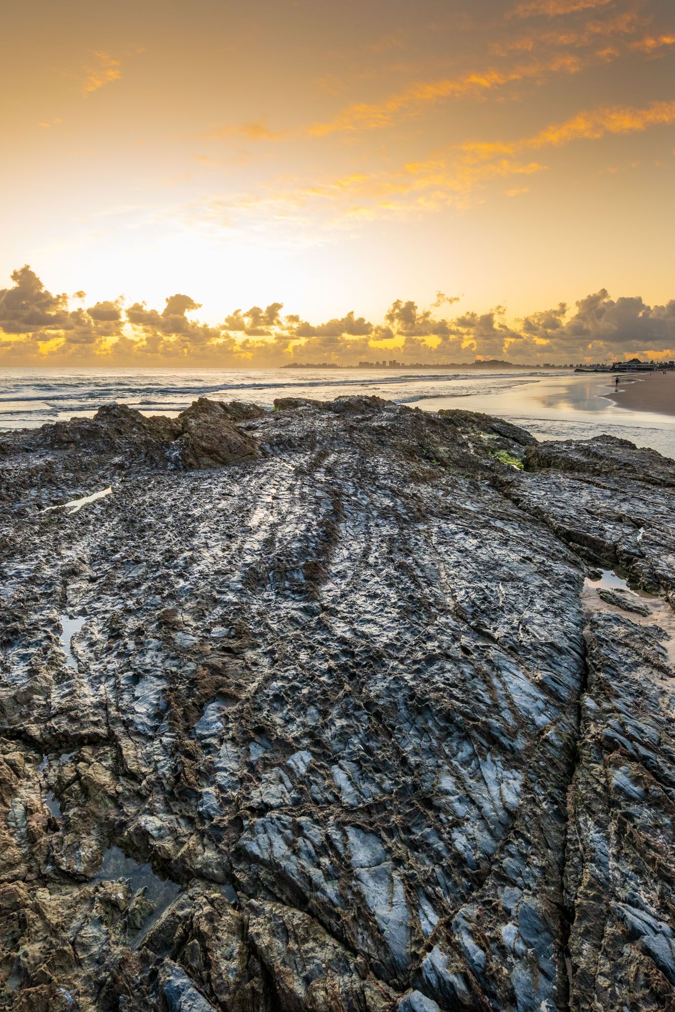 Rocky shoreline at golden sunrise with dramatic orange sky and clouds over calm ocean waters.