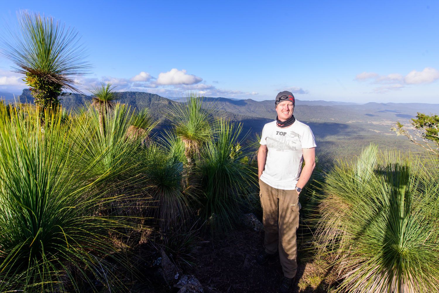 Man standing among grass trees on mountain summit with panoramic valley views under blue sky.