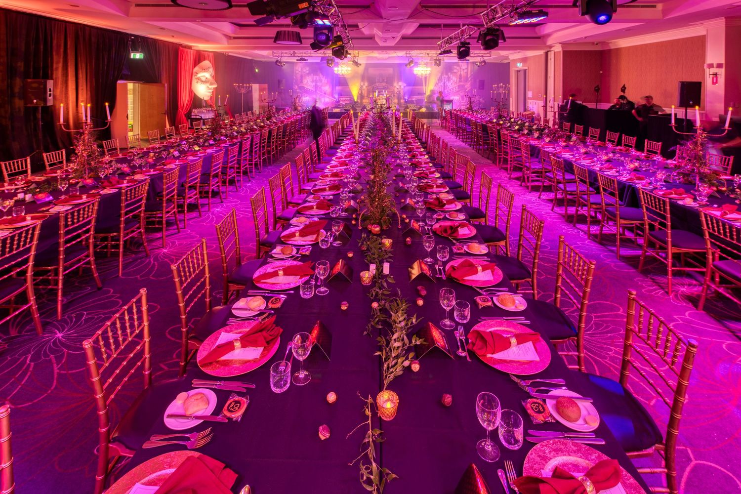 Elegant gala dinner banquet hall with long tables, gold chairs, pink floral centerpieces and vibrant purple lighting.