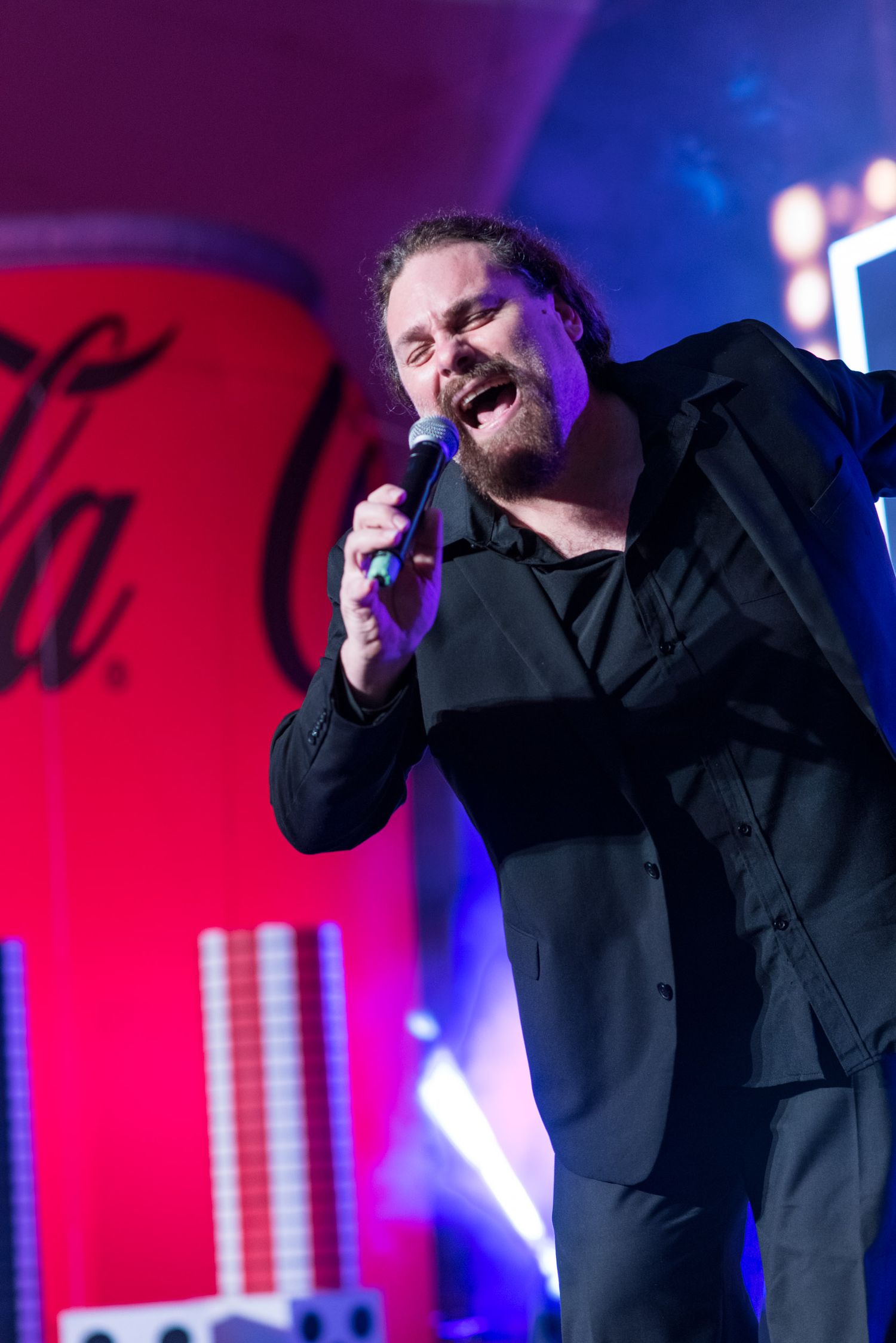 Male singer in black suit performing on stage with Coca-Cola branded backdrop and colorful stage lighting.