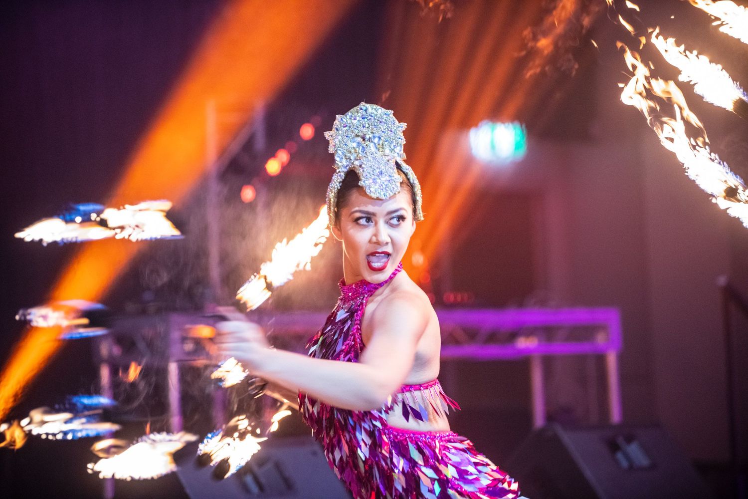 Dynamic female fire performer in sequined costume spinning fire props on stage with dramatic orange lighting.
