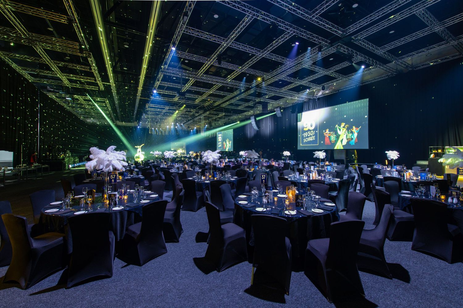Large gala dinner venue with green laser lights, black tables, white feathers, and projection screens.