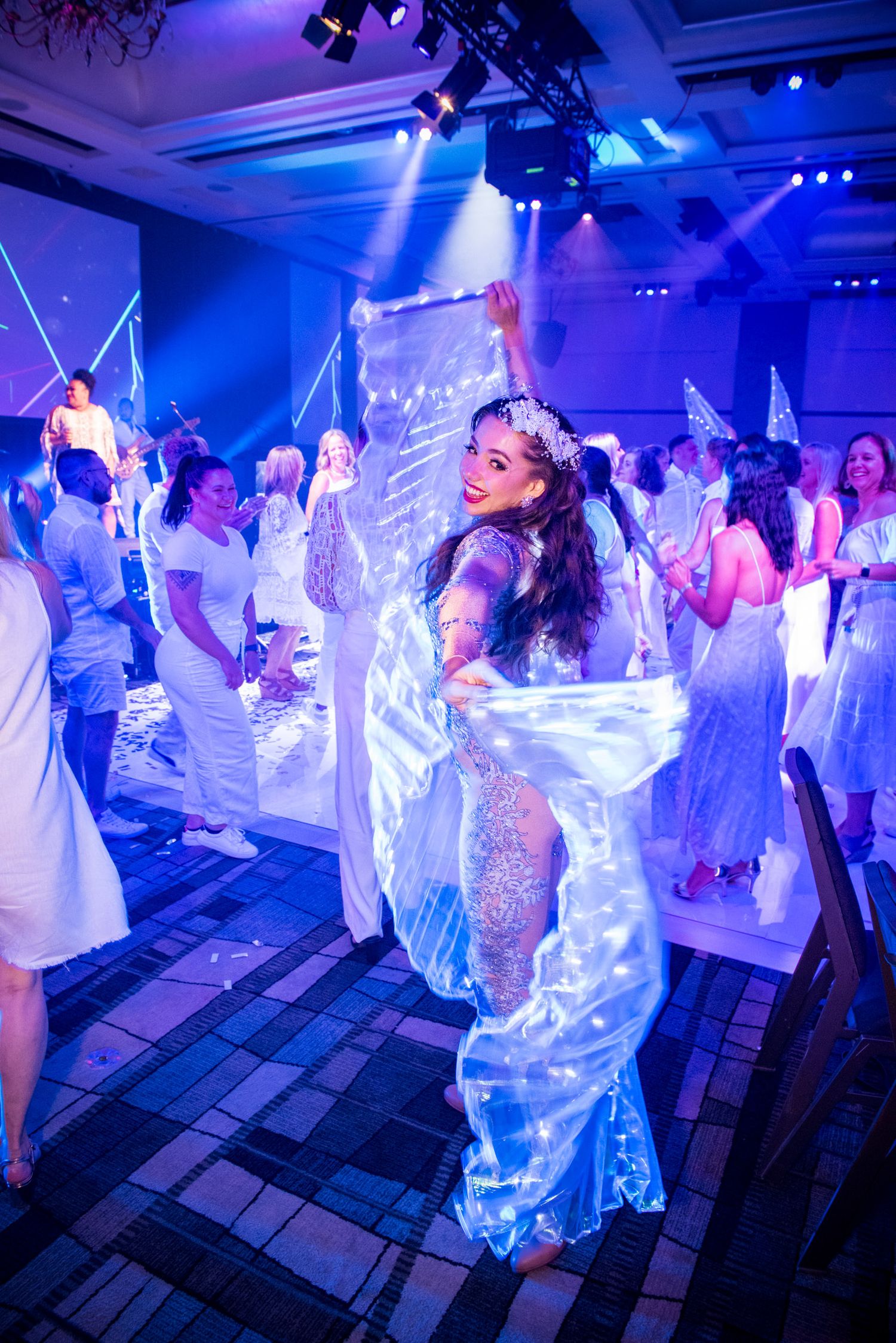 Woman in white dress dancing at an all-white themed corporate gala event with blue stage lighting and confetti.