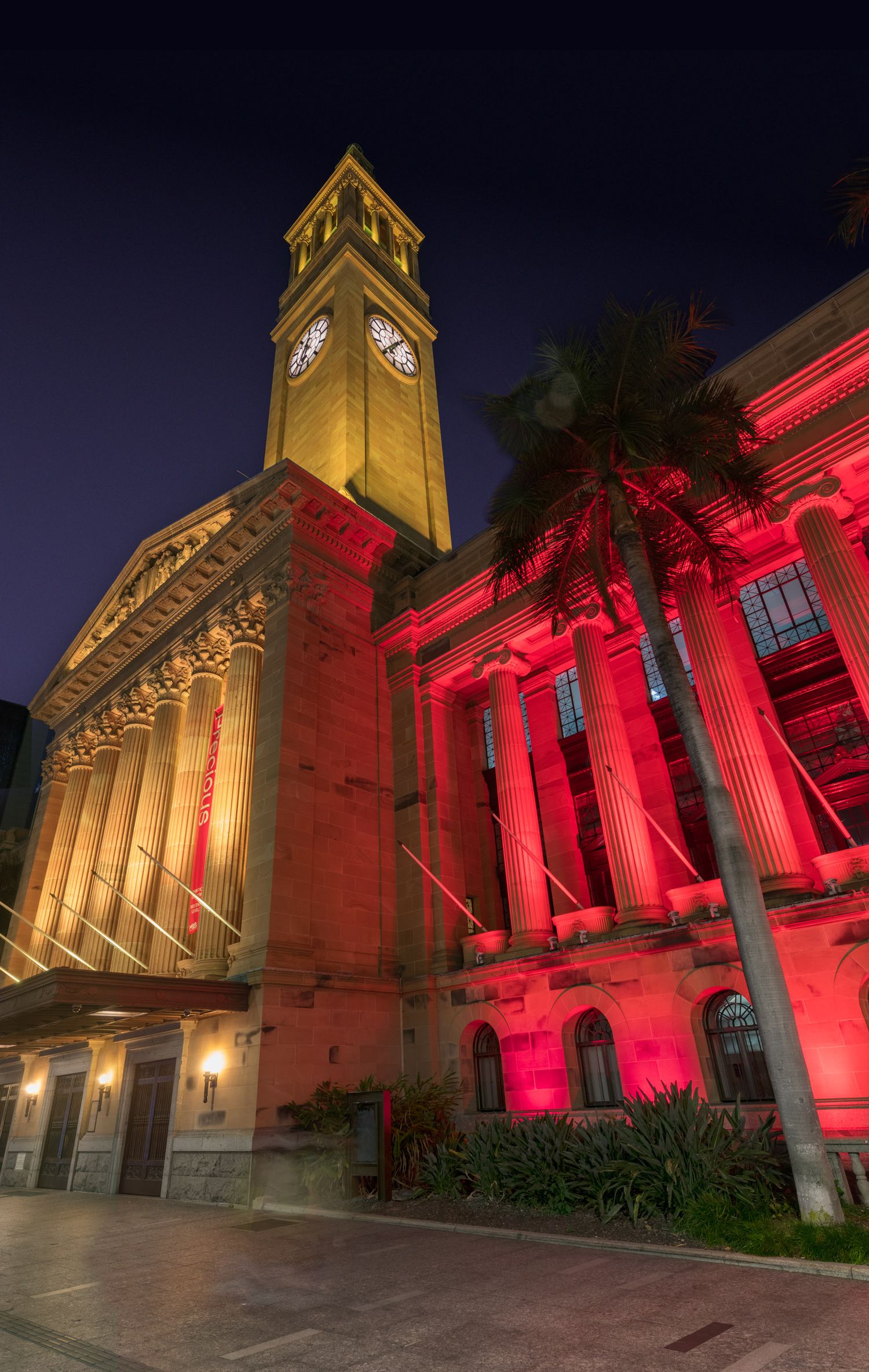 Brisbane City Hall clock tower dramatically lit in red and gold lights at night with palm trees in foreground.
