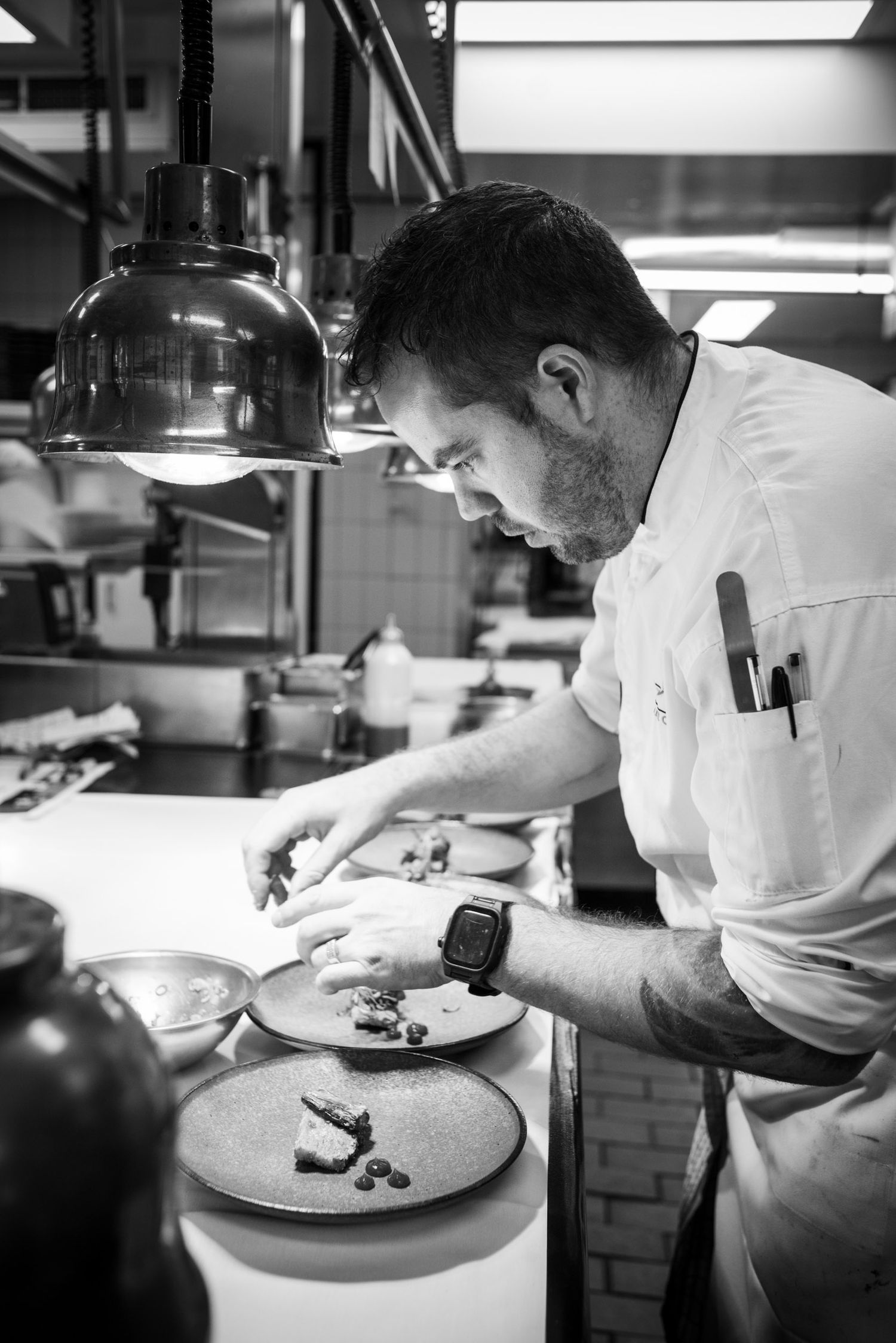 Black and white photo of focused chef plating dishes under kitchen heat lamps.