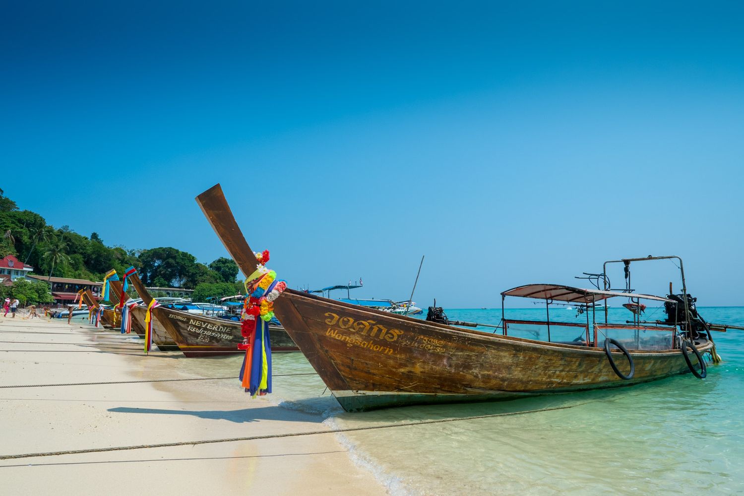 Colorfully decorated traditional Thai longtail boats lined up along a white sandy beach under a clear blue sky.