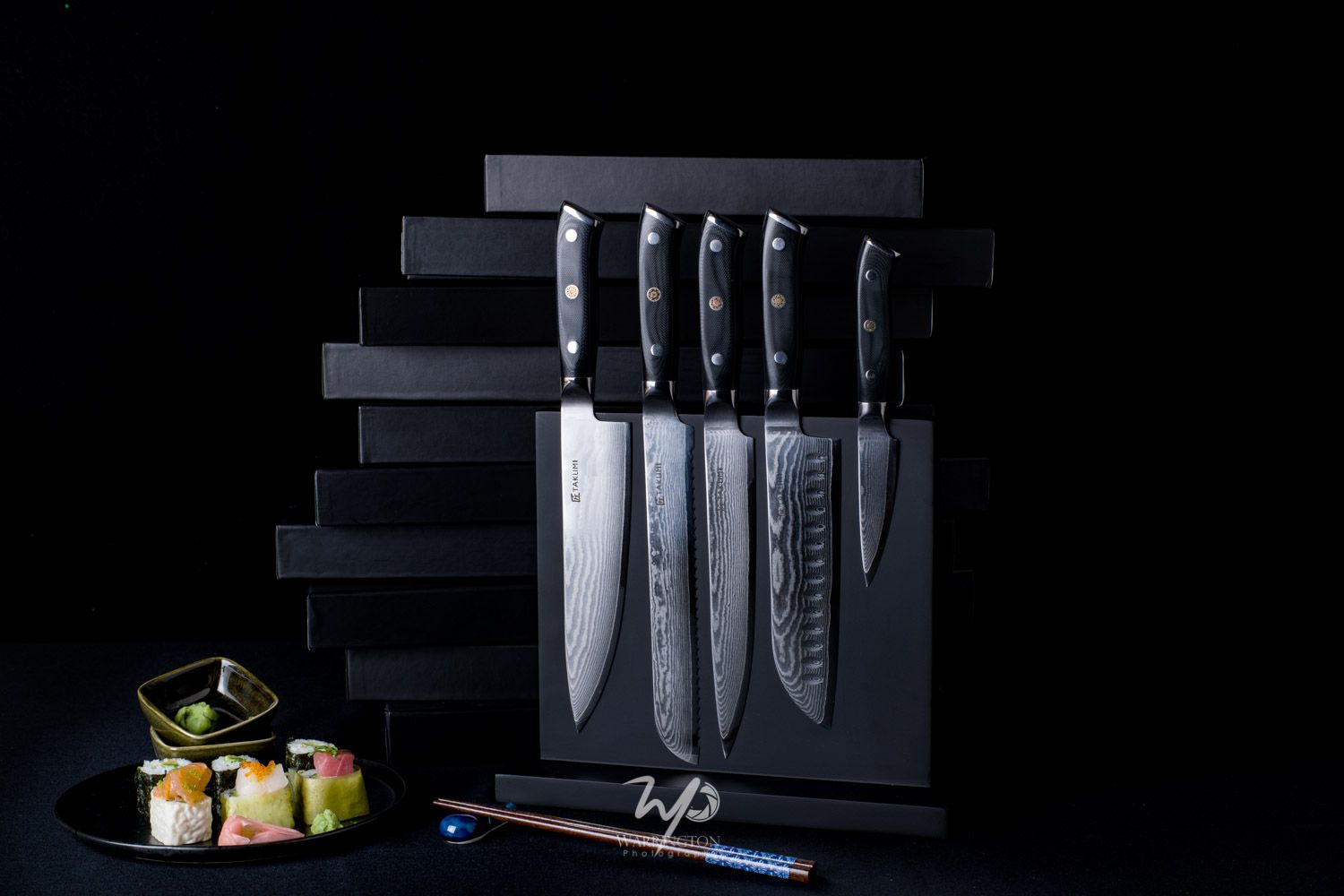 Set of five Damascus steel kitchen knives displayed on a black magnetic knife block with sushi in foreground.