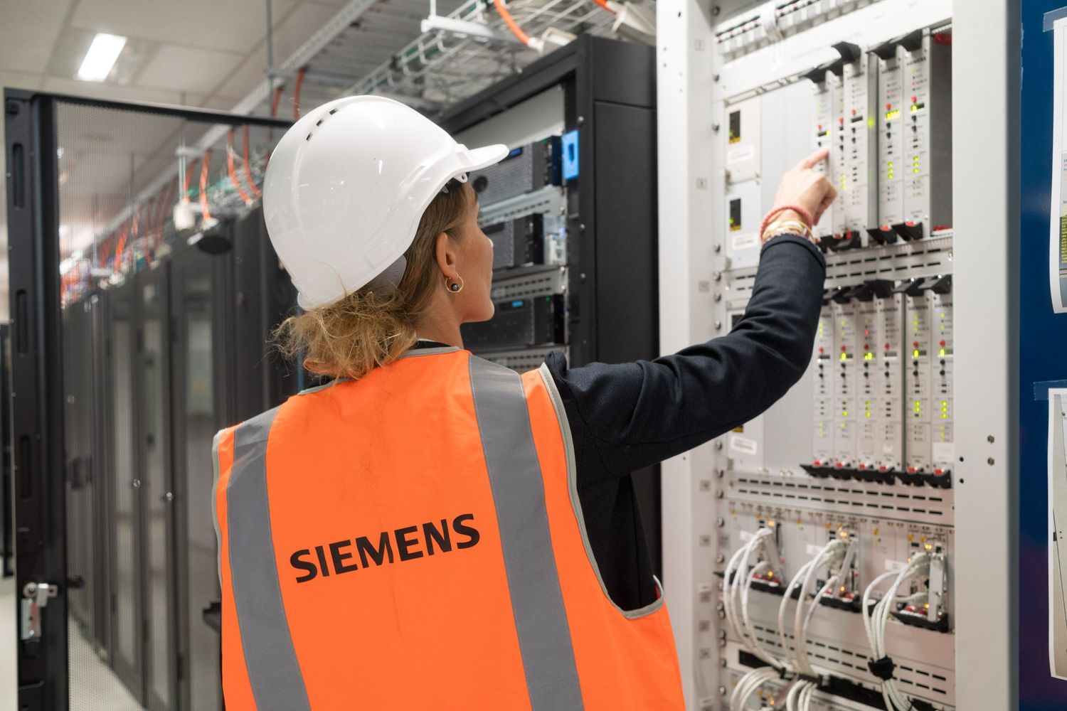 Female Siemens engineer in orange safety vest and white hard hat works on server equipment.