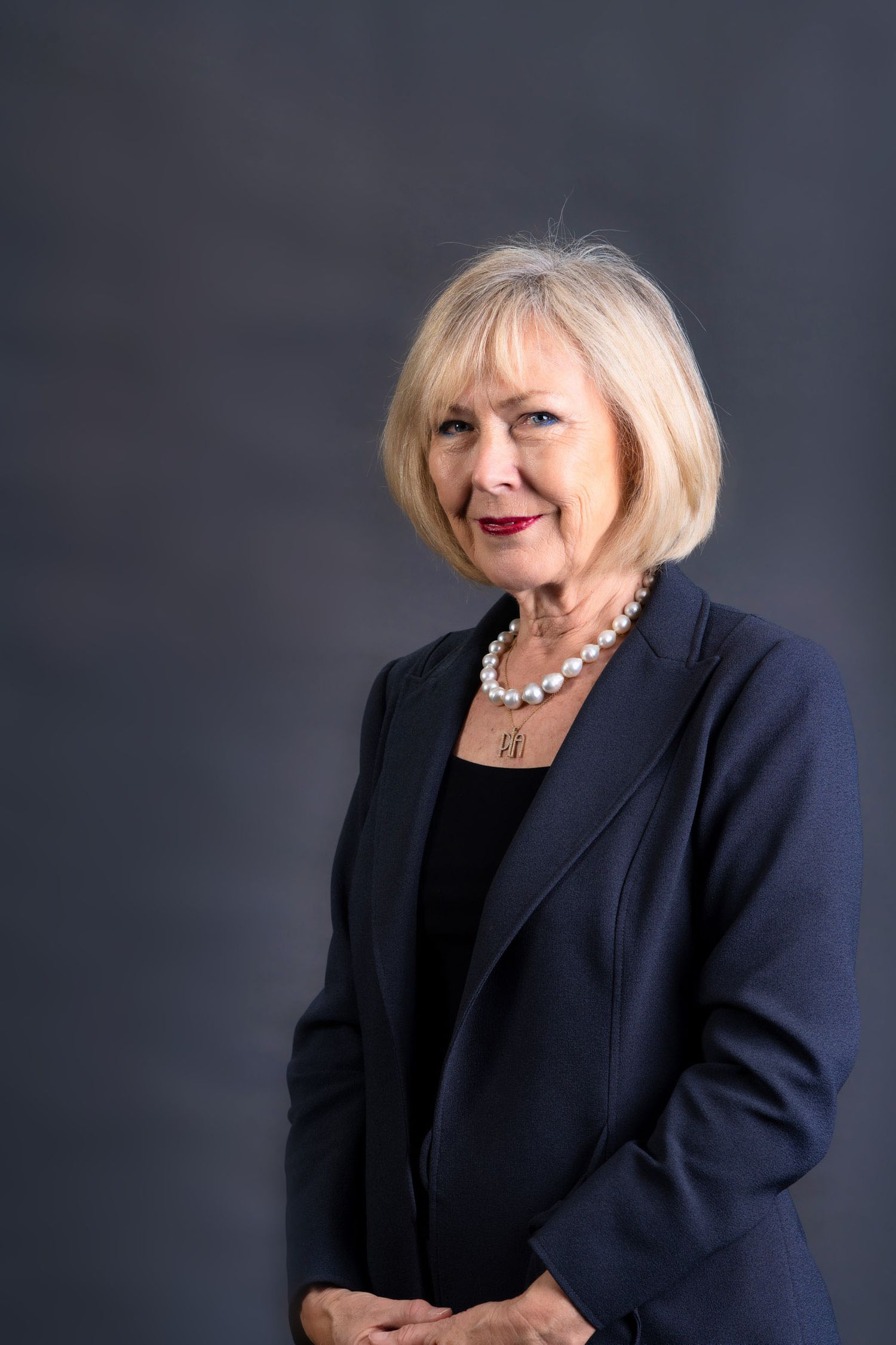 Elegant older woman with blonde hair wearing a navy blazer and pearl necklace smiling.