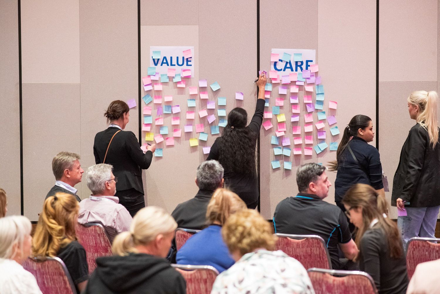 Conference attendees post colorful sticky notes on VALUE and CARE boards during a workshop activity.