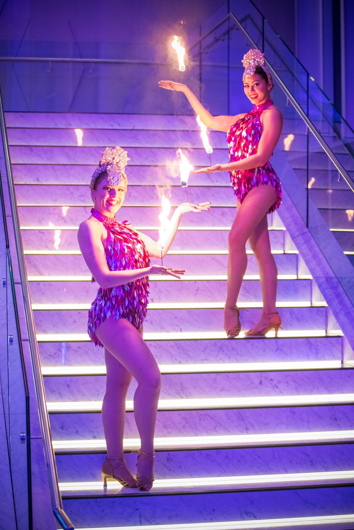 Two female fire performers in sequined costumes posing on purple-lit marble stairs.