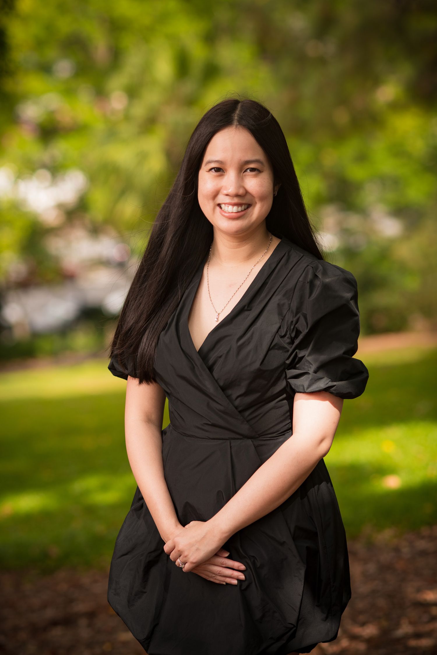 Young Asian woman smiling in a black dress posing outdoors in a lush green garden setting.