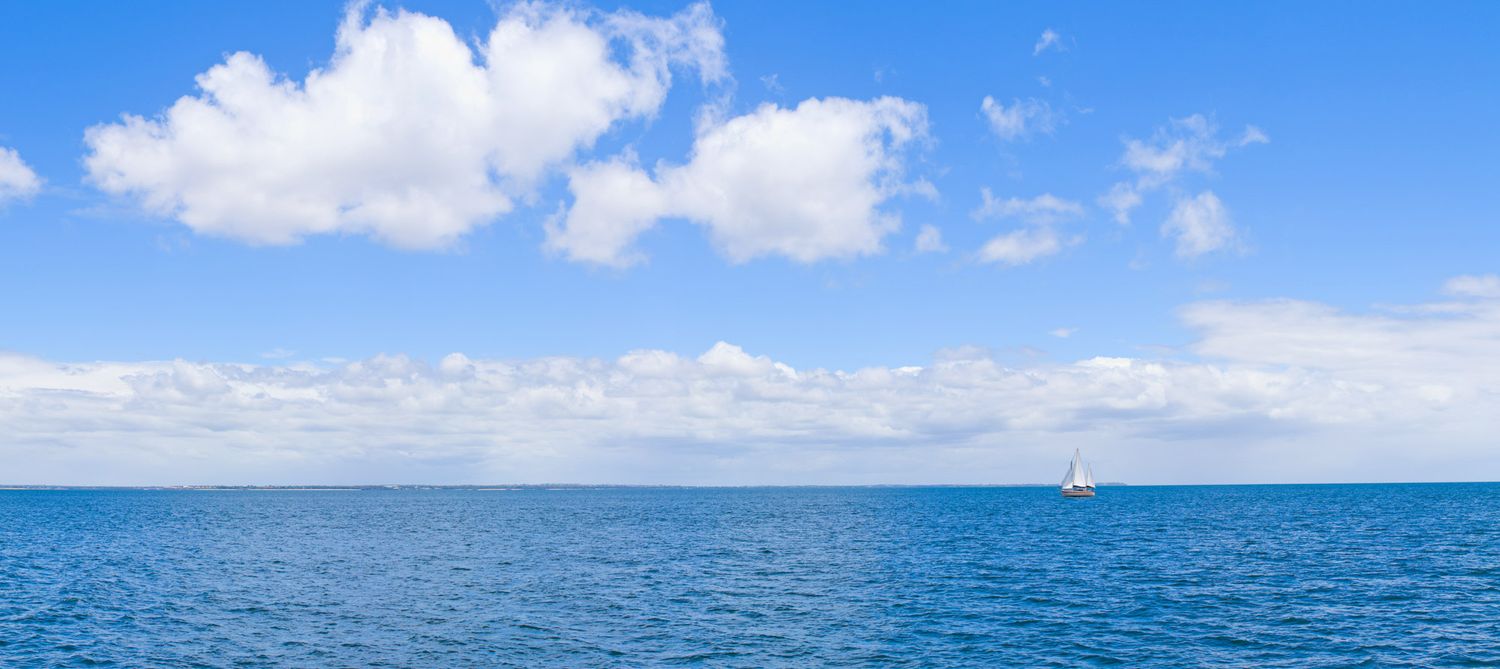 A sailboat glides across calm blue ocean waters under a bright sky with fluffy white clouds on the horizon.