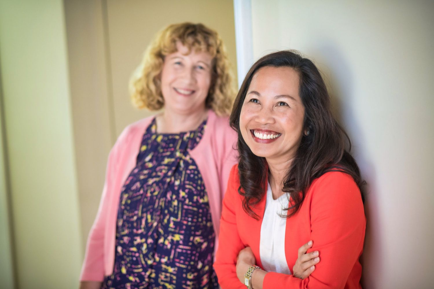 Two smiling professional women posing together indoors, one in pink cardigan and one in red blazer.