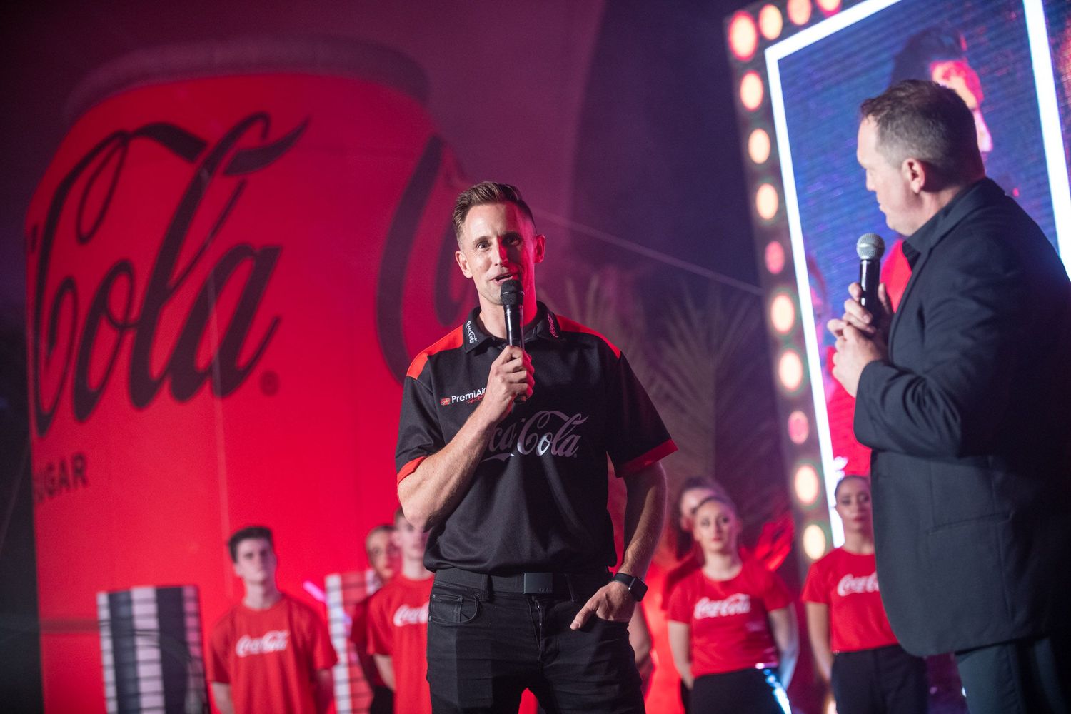 Two men speak on stage at a Coca-Cola branded evening event with red lighting and a large can prop.