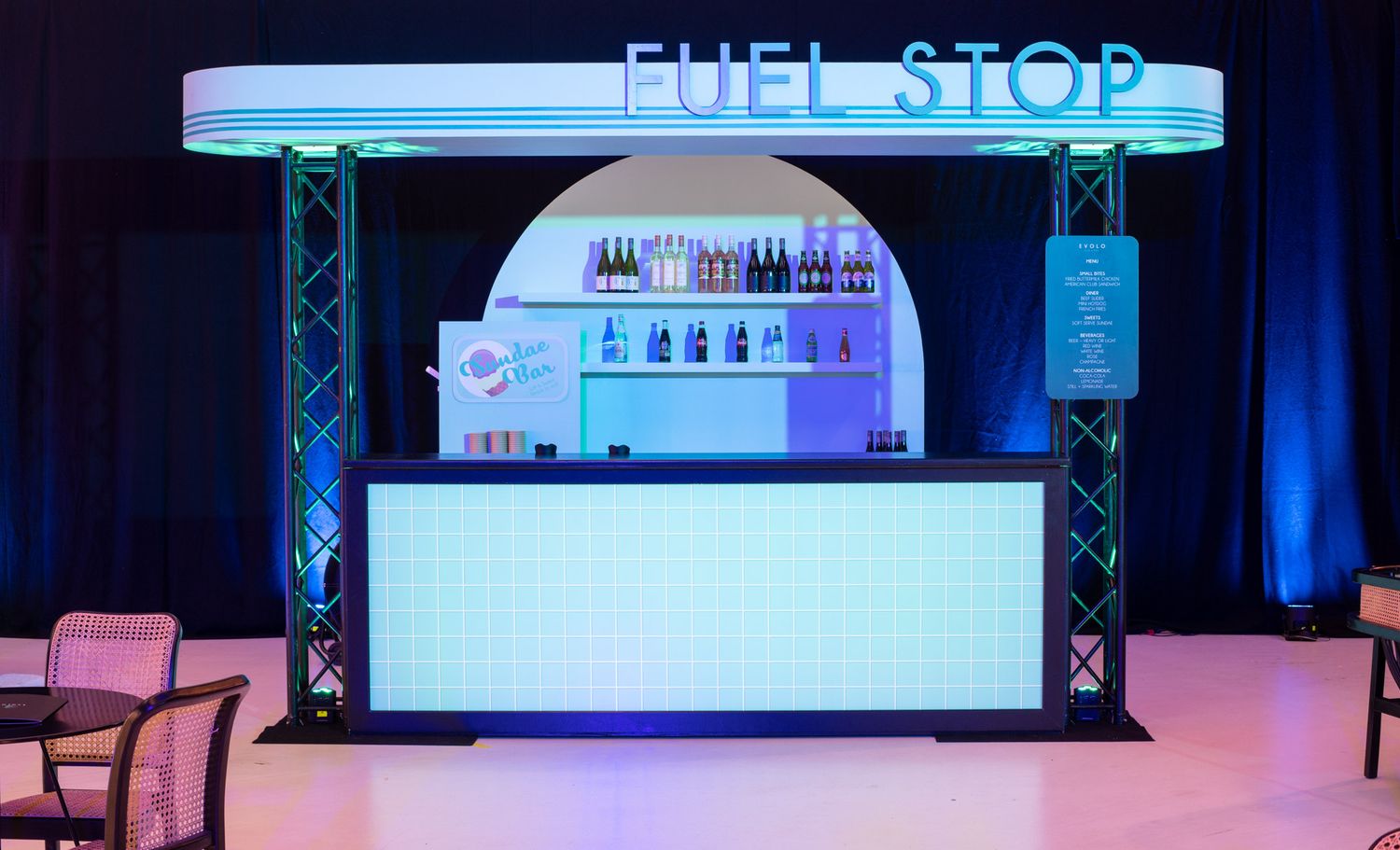 A themed bar setup called Fuel Stop with LED panels, bottle displays, and teal lighting at an event.