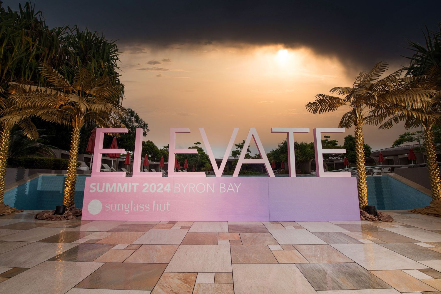 Large pink and white ELEVATE Summit 2024 sign beside a pool with palm trees at dusk.