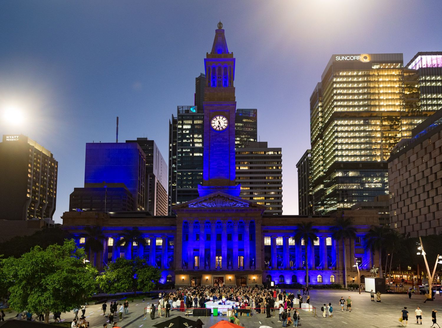 Brisbane City Hall illuminated in blue lights at dusk surrounded by city skyscrapers.