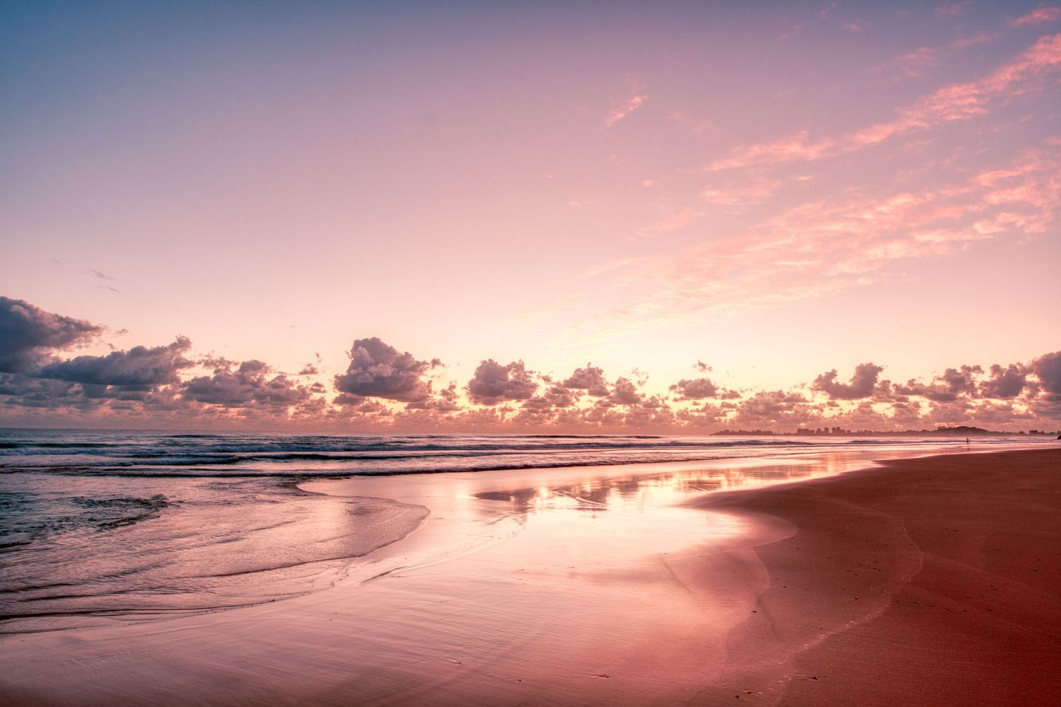 Serene pink sunset reflecting on wet sandy beach with gentle waves and clouds.