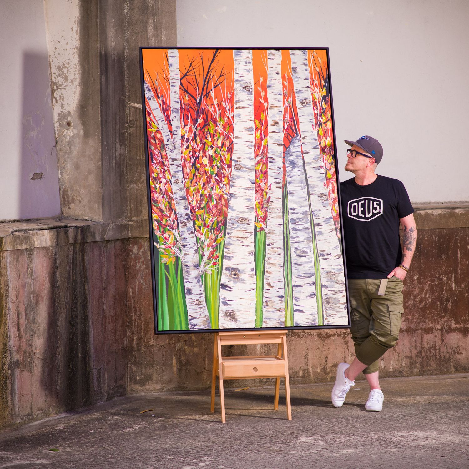 Artist standing beside a large colorful birch tree painting on an easel in an urban setting.