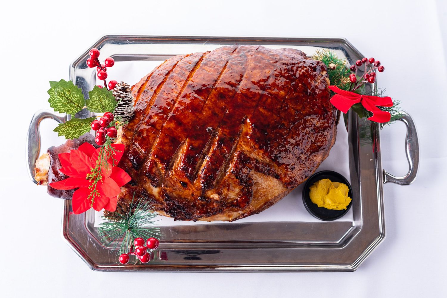 Glazed Christmas ham on silver tray decorated with poinsettia, holly, and mustard sauce.