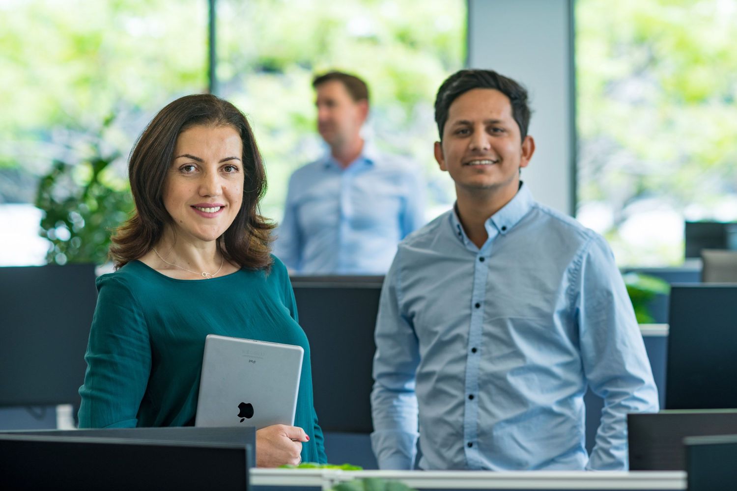 Professional woman holding tablet and smiling man stand in modern office with colleague working in background.