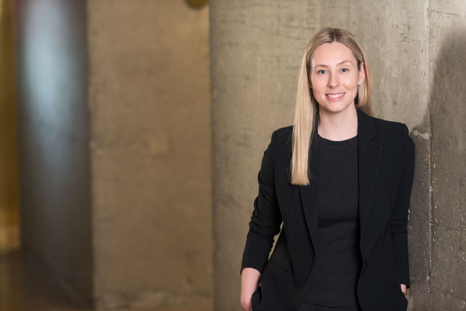 Professional woman in a black blazer leaning against a concrete wall smiling confidently.
