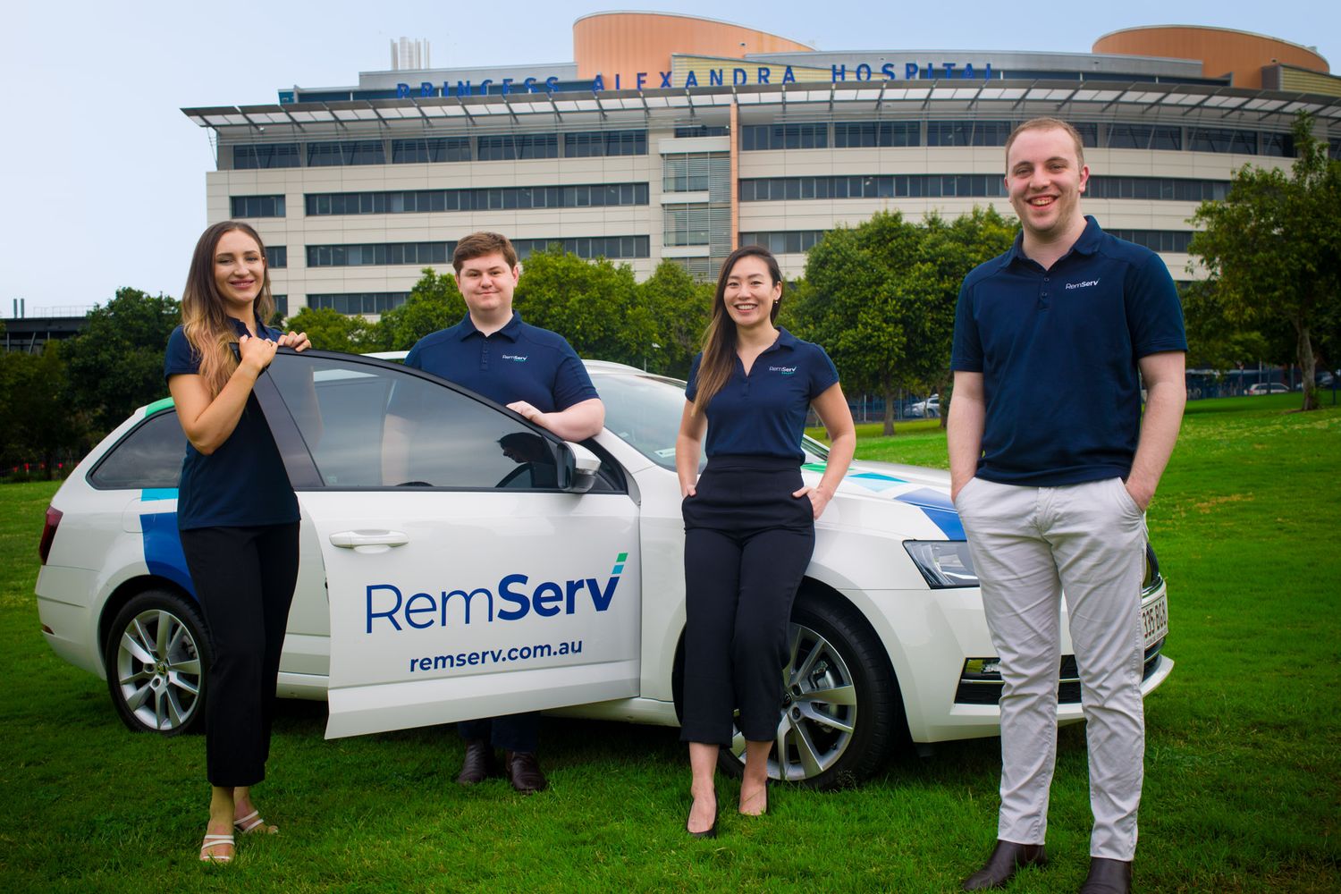 Four RemServ employees smiling beside branded company car outside Alexandra Hospital on sunny green lawn.