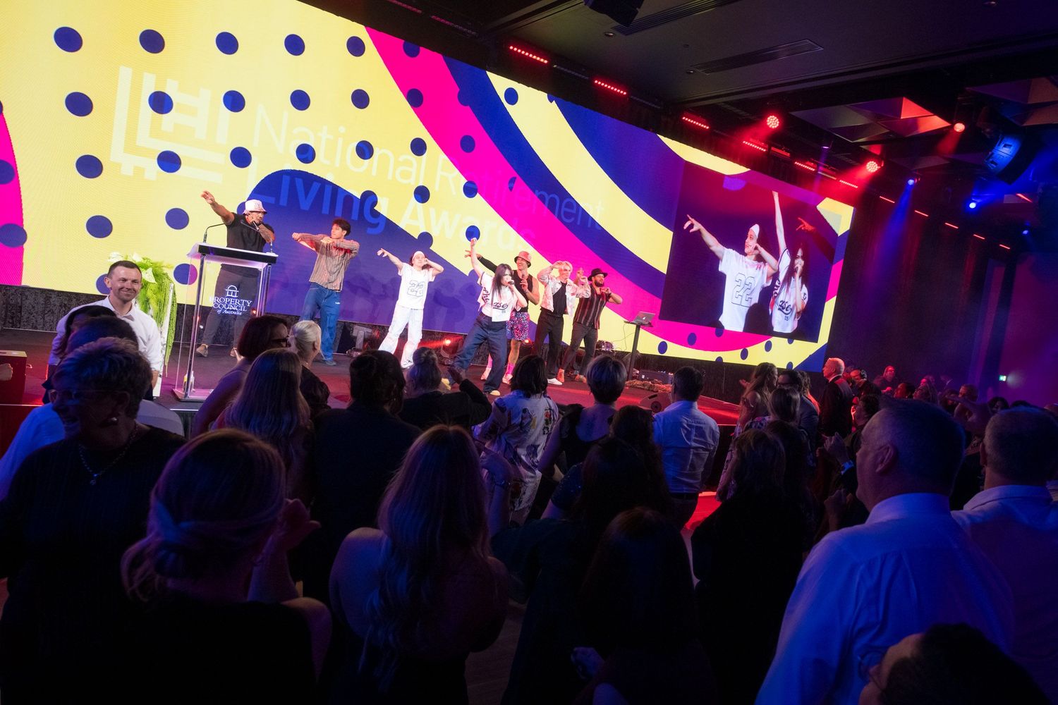 Energetic performers on colorful stage entertain large crowd at corporate awards event.