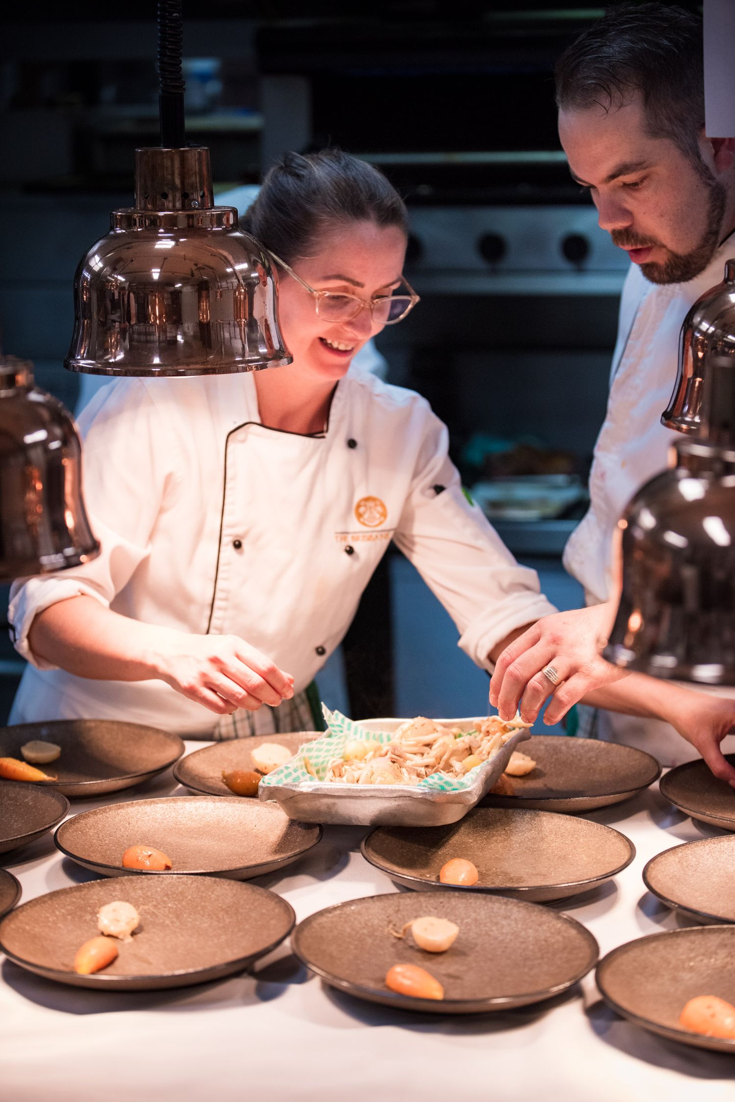 Two chefs in white uniforms plating gourmet dishes under warm lights in a restaurant kitchen.