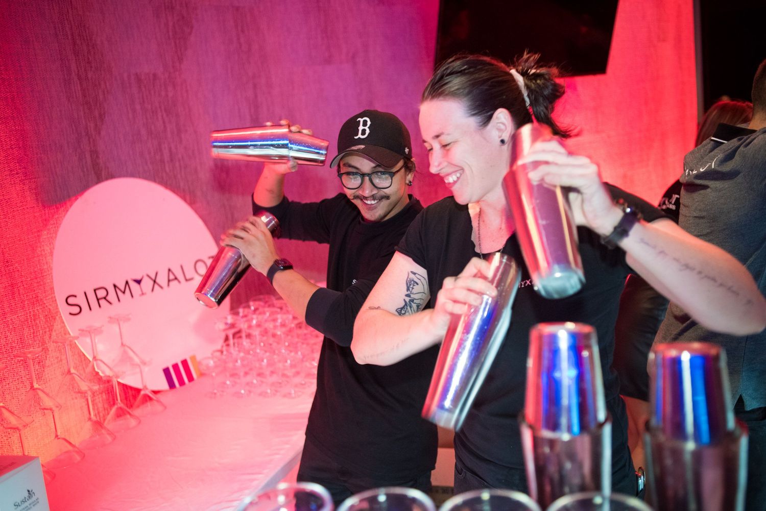 Two bartenders in black caps enthusiastically shaking cocktails at a vibrant neon-lit bar event.