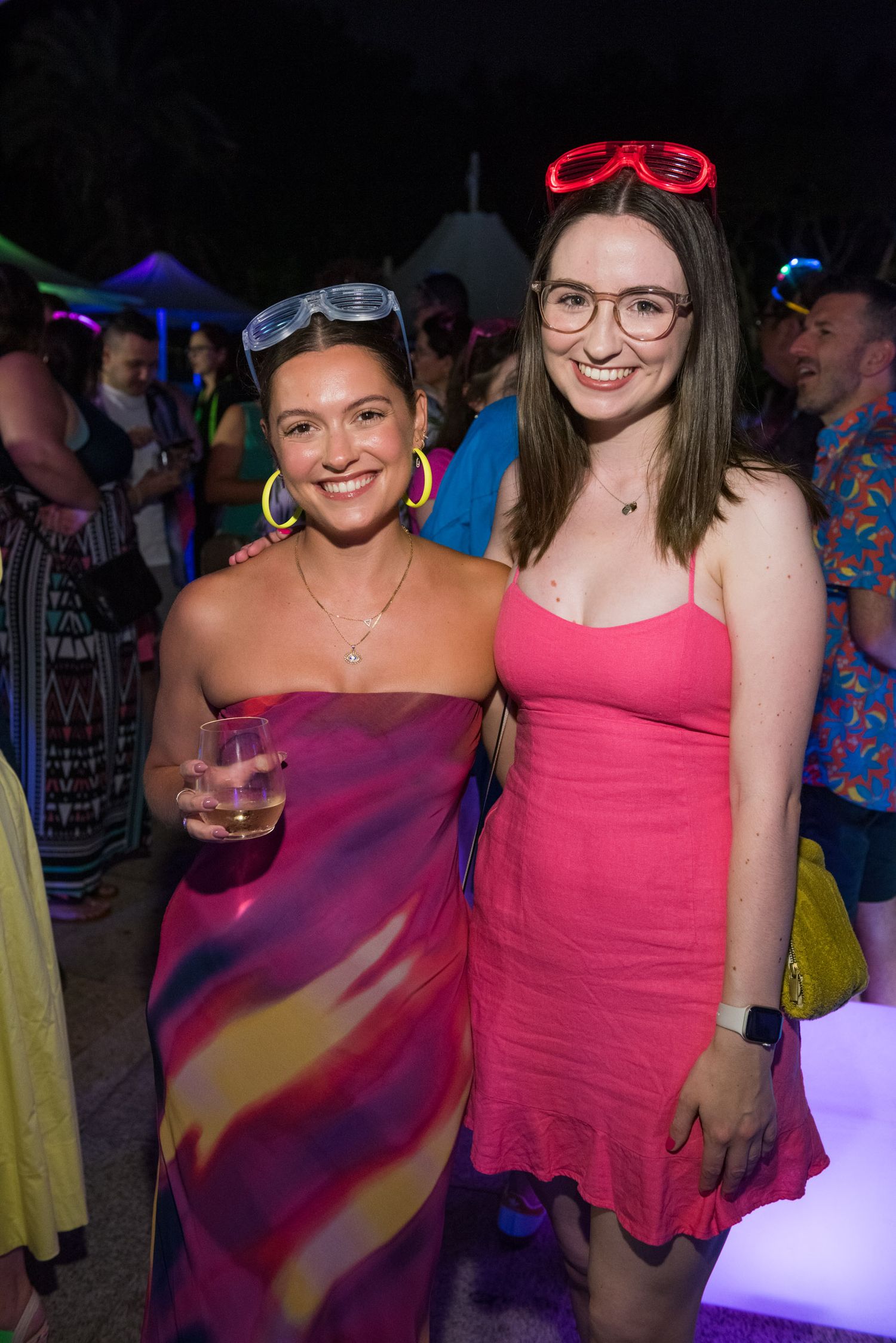 Two smiling women in colorful dresses posing together at a vibrant outdoor nighttime party event.