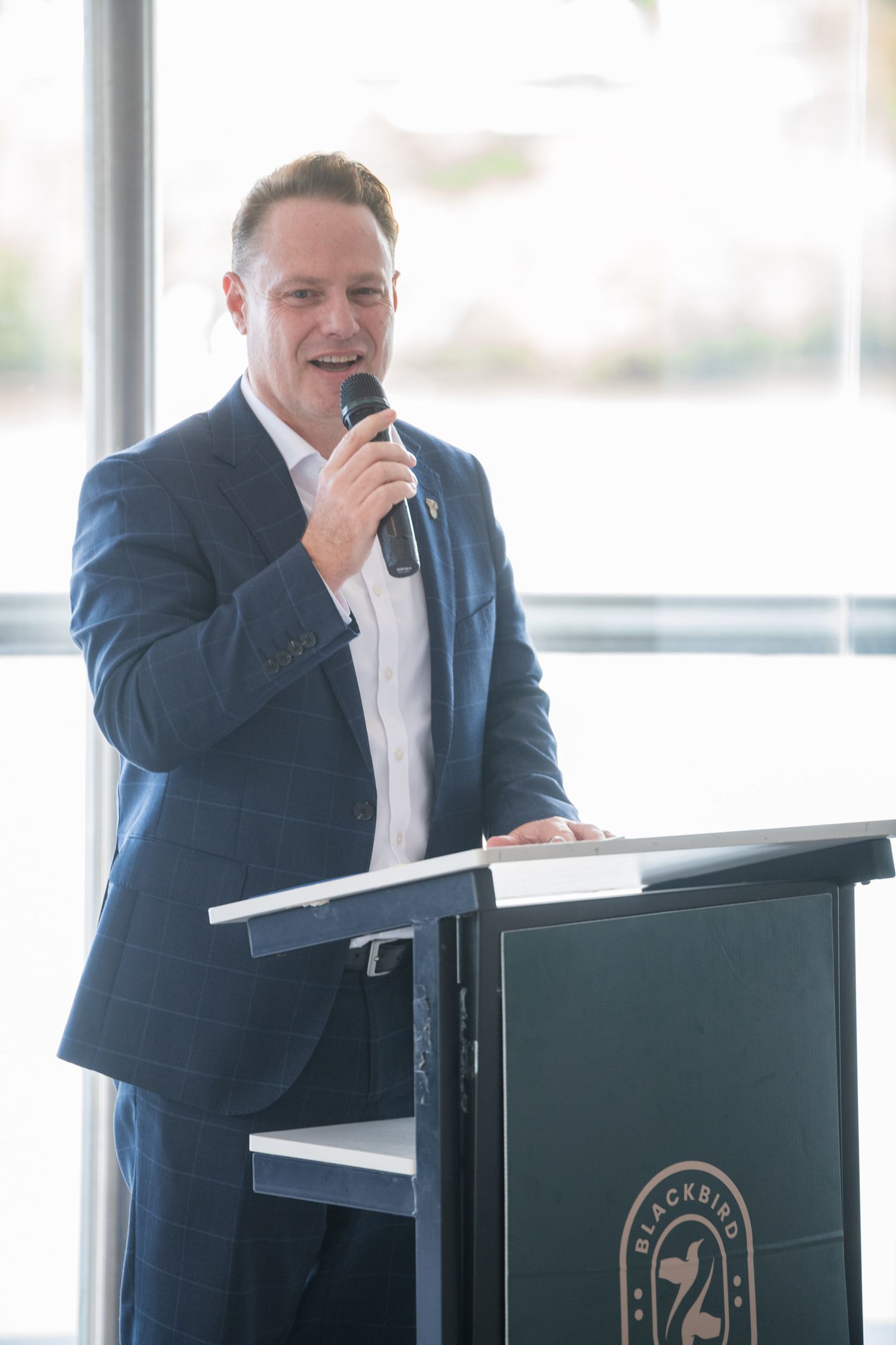 Confident man in blue blazer speaking at a Blackbird-branded podium holding a microphone.