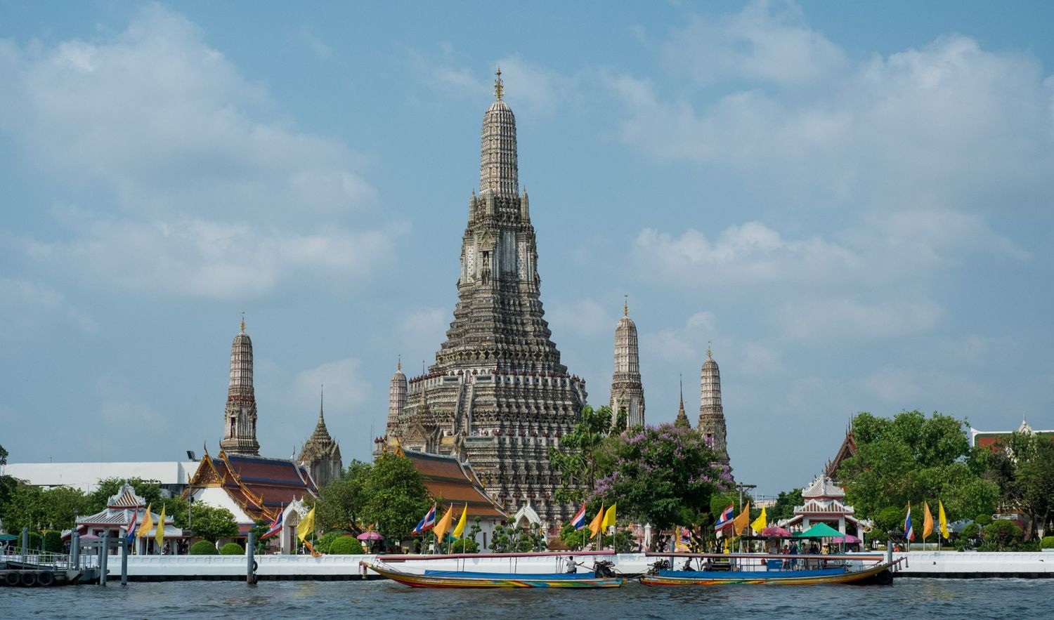 Iconic Wat Arun temple rising above the Chao Phraya River in Bangkok with colorful boats passing by.