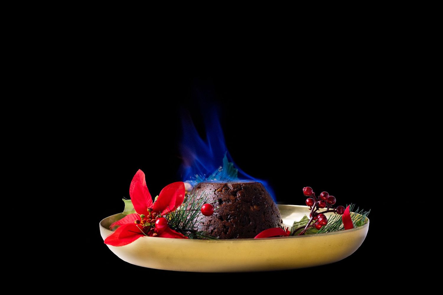 Traditional Christmas pudding flambeed with blue brandy flames, decorated with holly and poinsettia.